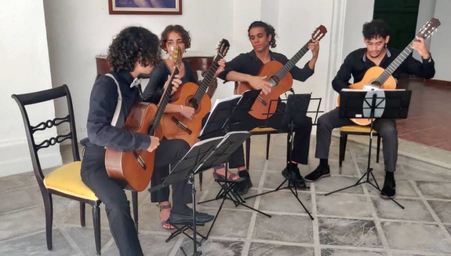 Cuarteto de músicos jóvenes tocando guitarras, vestidos de negro, en un ambiente interior con sillas y partituras.
