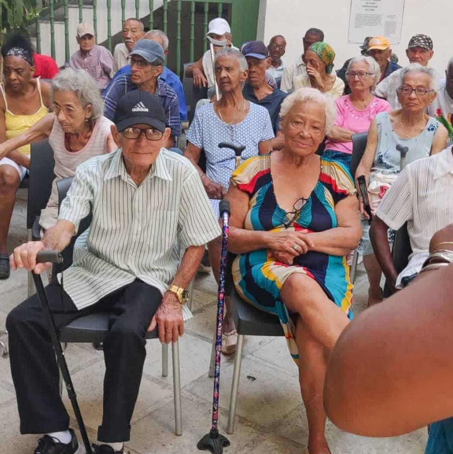 Grupo de personas mayores sentadas en sillas, participando en un evento o actividad social al aire libre.