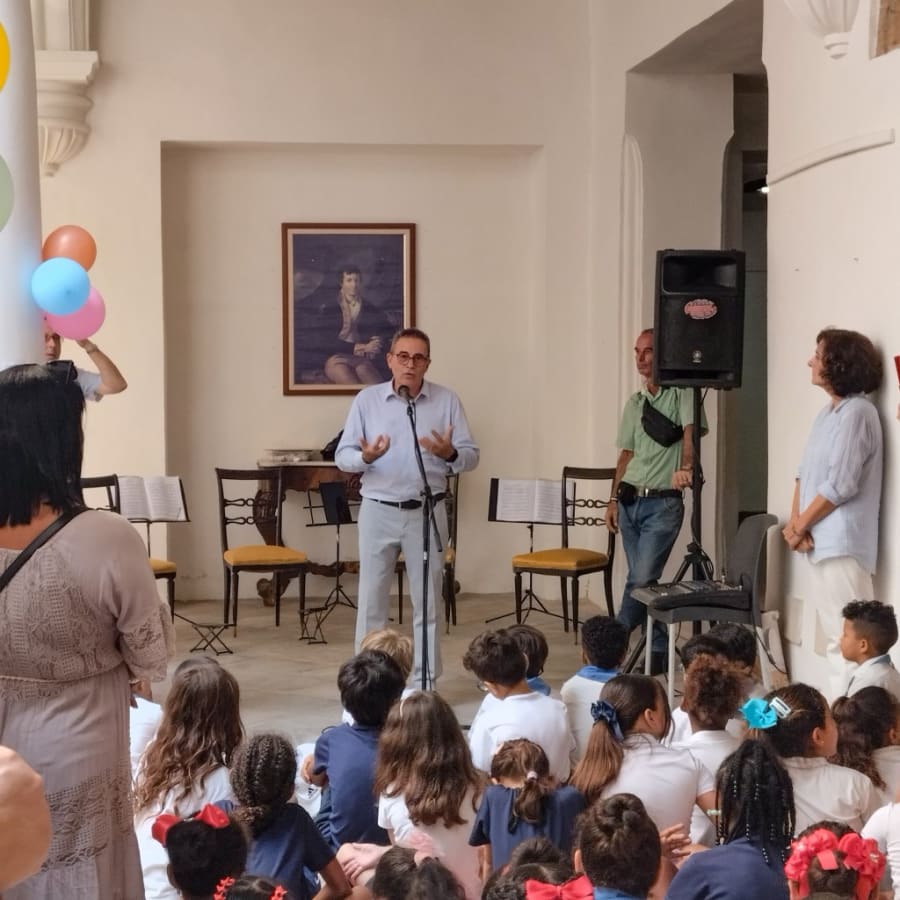 Una persona hablando en un micrófono frente a un grupo de niños sentados en el suelo. El ambiente es festivo, con globos de colores en el fondo y una pintura en la pared.