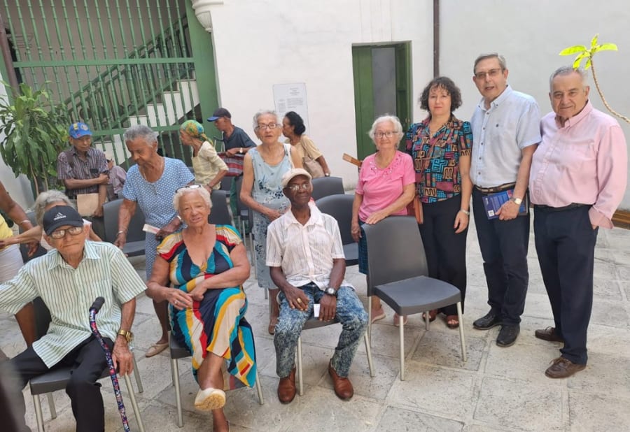 Grupo de personas mayores y académicos reunidos en un evento en Cuba, con un fondo que incluye plantas y sillas. Algunos participantes están sentados, mientras que otros se encuentran de pie en un entorno interior.