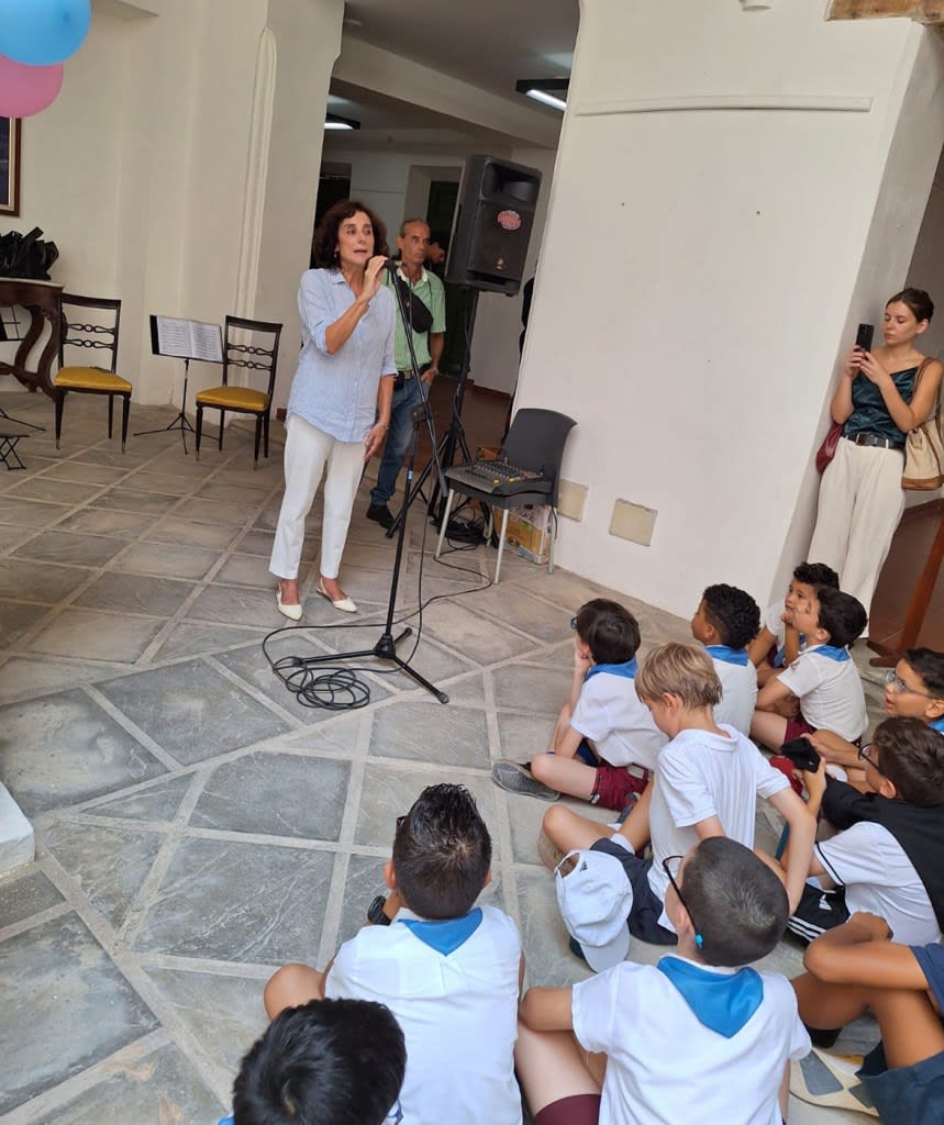 Una mujer habla en un micrófono frente a un grupo de niños sentados en el suelo, durante un evento conmemorativo en Cuba.