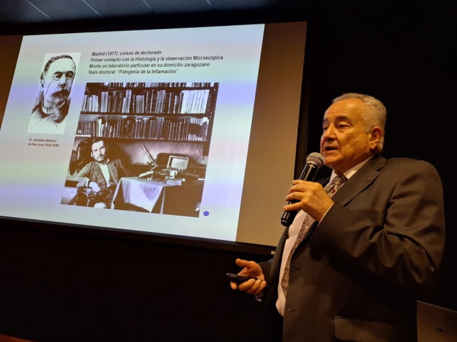 Un ponente presenta información sobre Santiago Ramón y Cajal en una conferencia, con imágenes históricas de Cajal y otros científicos en una pantalla de fondo.