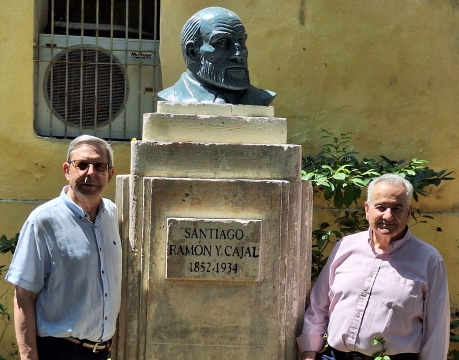Dos hombres posan junto a un busto de Santiago Ramón y Cajal, ubicado en un entorno verde. La escultura está en un pedestal con una placa que lleva su nombre y años de vida.