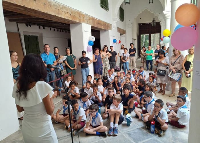 Un grupo de niños sentados en el suelo, atentos a una oradora en una sala, con personas de pie detrás de ellos, decoraciones coloridas en el fondo.