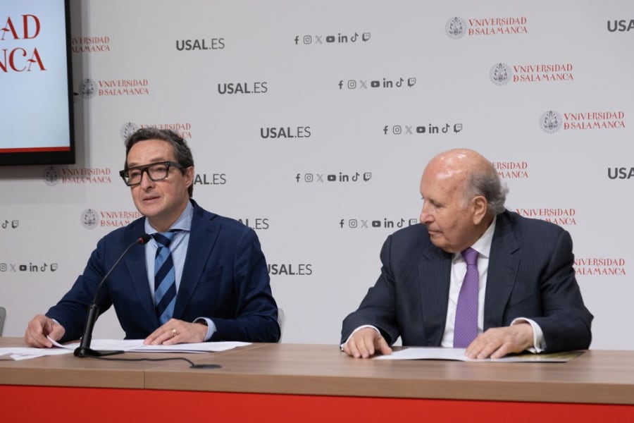 Dos hombres sentados en una mesa durante una rueda de prensa en la Universidad de Salamanca, con un fondo decorado con el logotipo de la universidad.