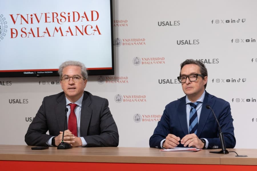 Dos hombres sentados en una mesa durante una rueda de prensa en la Universidad de Salamanca, con el logo de la universidad detrás de ellos.