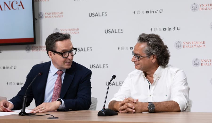 Dos hombres sentados en una mesa durante una rueda de prensa en la Universidad de Salamanca, con un logo en el fondo.