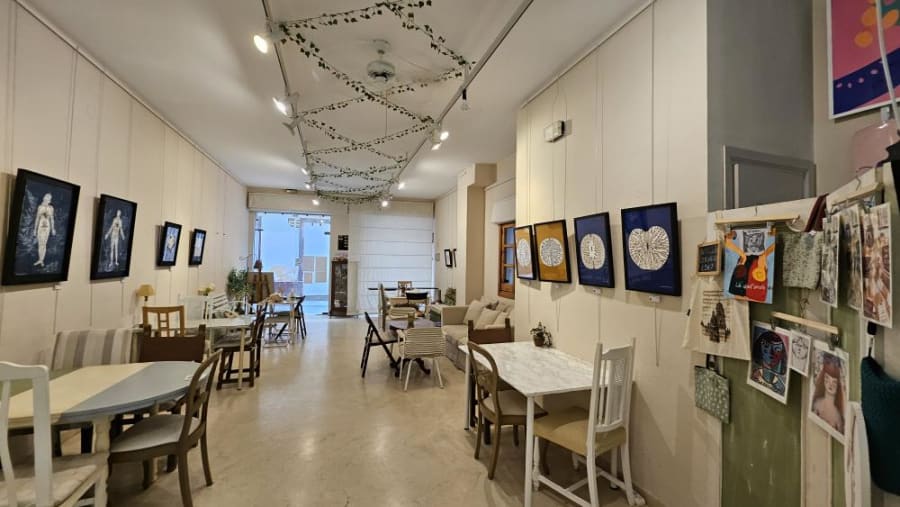 Interior de un espacio cultural con mesas y sillas dispuestas para visitantes, exhibiendo obras de arte enmarcadas en las paredes, en un ambiente luminoso y acogedor.