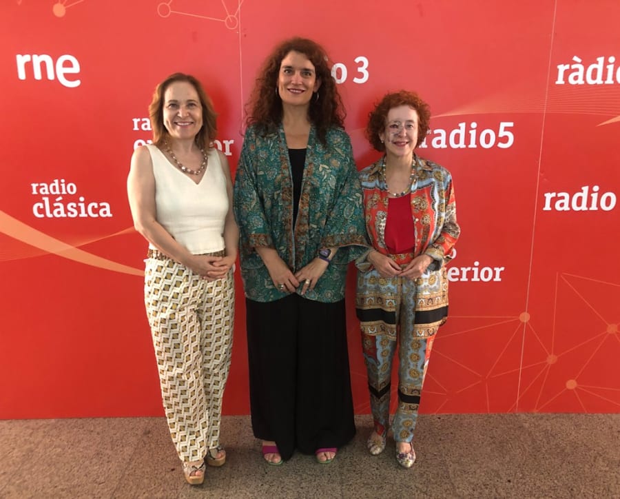 Tres mujeres sonrientes posan juntas frente a un fondo rojo con el logo de 