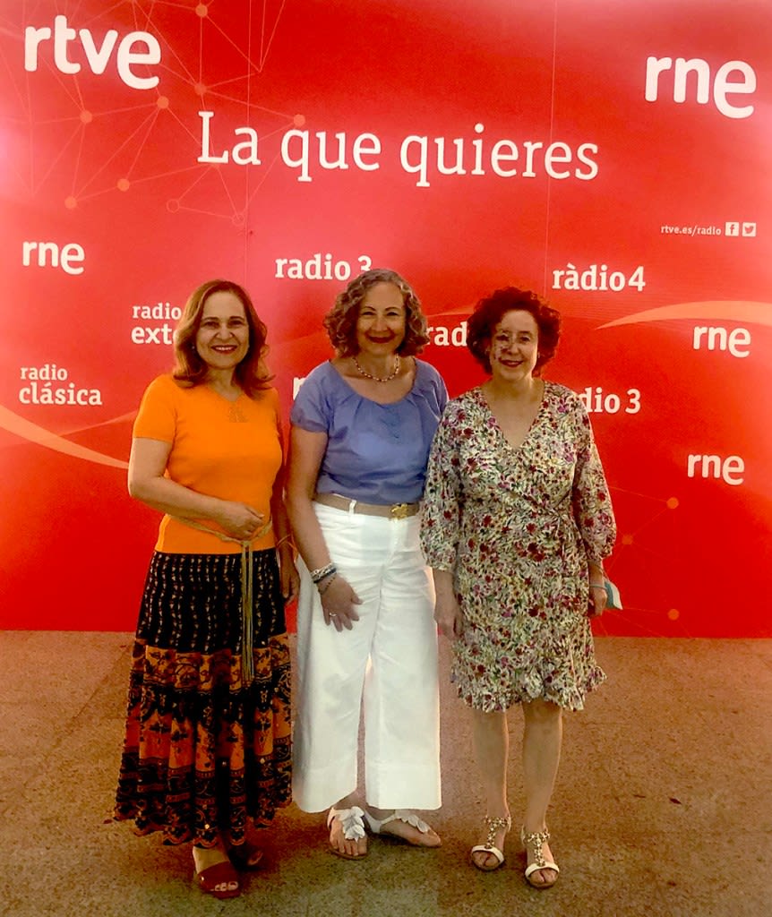 Tres mujeres posan frente a un fondo rojo con el logo de RTVE, mostrando un ambiente festivo y amistoso.
