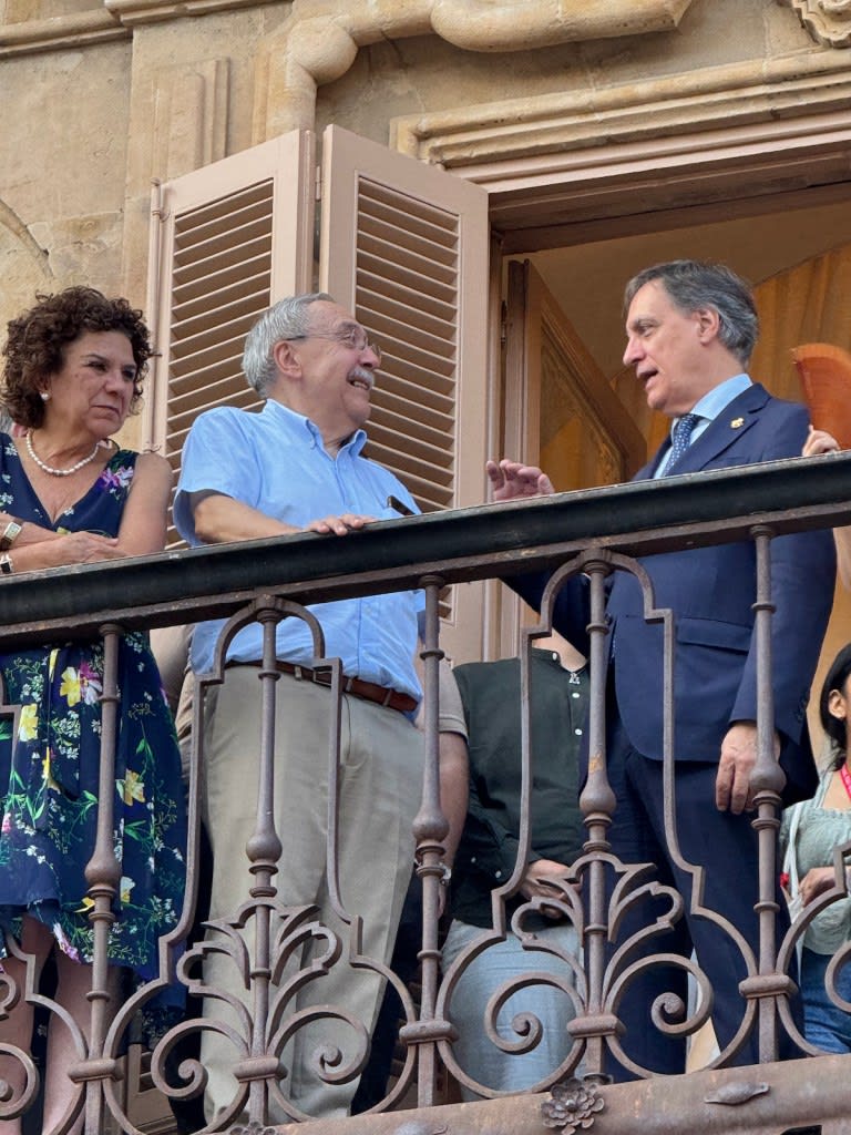 Tres personas conversando en un balcón con barandilla decorativa, rodeadas de un ambiente arquitectónico clásico.