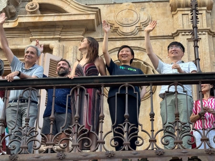 Grupo de personas en un balcón, saludando y sonriendo. Presentan una variedad de expresiones de alegría y camaradería en un evento al aire libre.