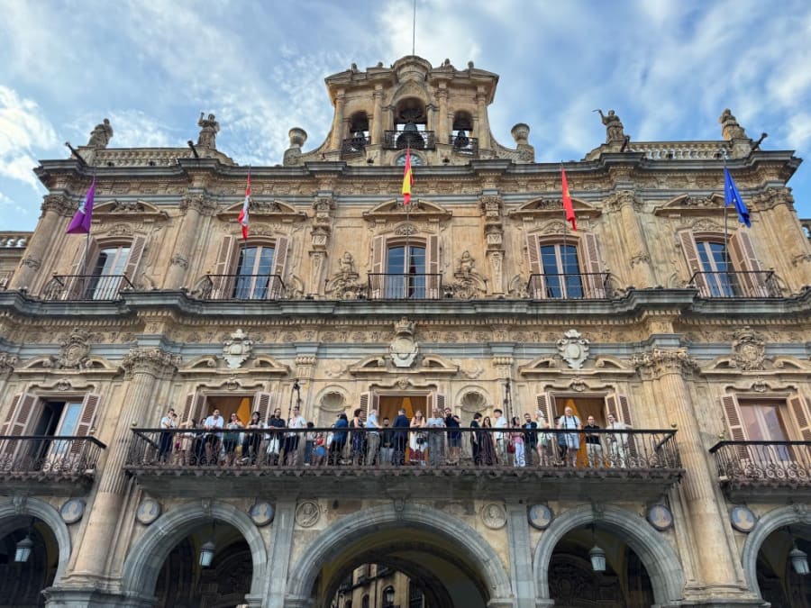 Vista de la fachada del edificio histórico con varias personas asomadas al balcón, adornado con banderas de España y de diversas comunidades autónomas.