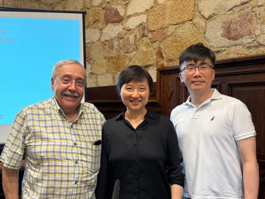 Tres científicos expertos posando juntos en un evento académico, con un fondo de pared de piedra y una presentación en pantalla detrás.