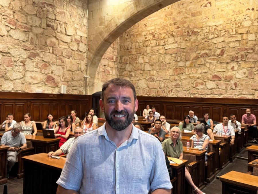 Retrato de un hombre sonriente frente a un grupo de estudiantes en un aula con paredes de piedra, durante el curso de Biofísica en la Universidad de Salamanca.