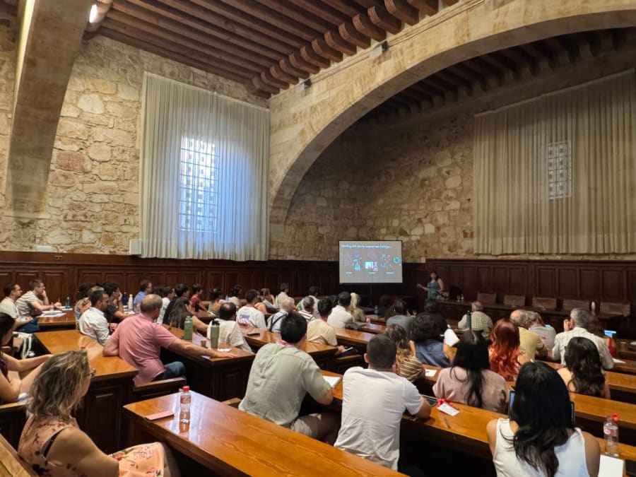 Un grupo de estudiantes y profesores asiste a una conferencia en un aula con paredes de piedra, donde se presenta información sobre biofísica. Se pueden ver sillas de madera y un proyector mostrando diapositivas.