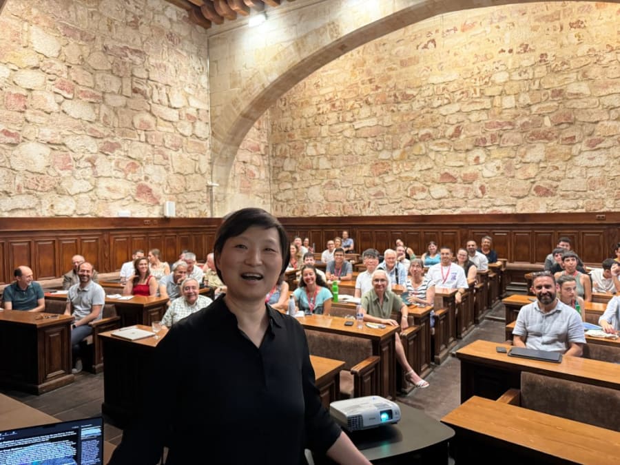 Imagen de una conferencia en la Universidad de Salamanca, con un grupo de personas sentadas en un aula histórica y una presentadora en primer plano sonriendo.