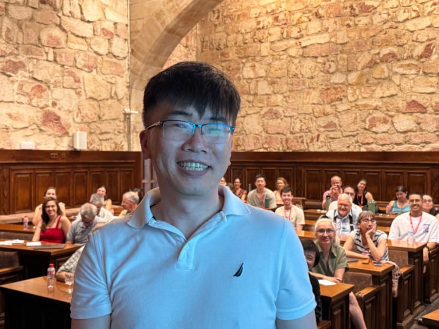 Un participante sonriente en un aula con barandillas de madera, rodeado de otros asistentes al curso de Biofísica en la Universidad de Salamanca.