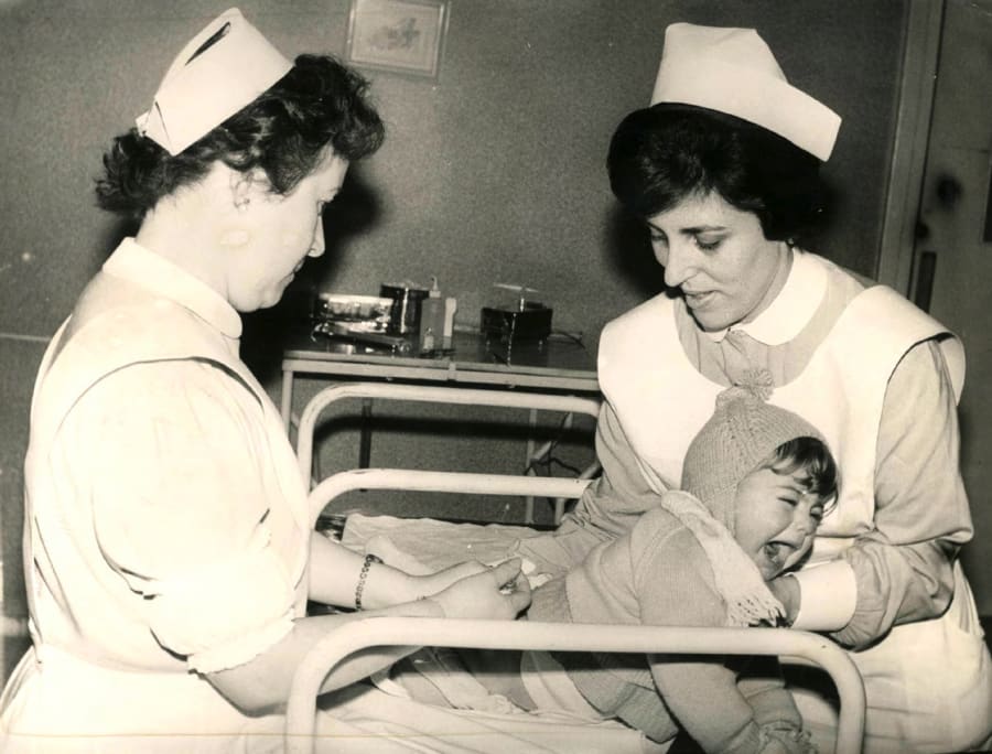 Dos enfermeras administrando una vacuna a un niño que llora en un hospital, en un ambiente clínico de la década de 1960.