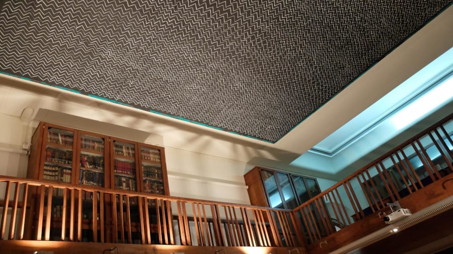 Vista del interior de una biblioteca, mostrando estanterías de madera con libros antiguos y un techo decorado con un diseño en zigzag en color negro y blanco.