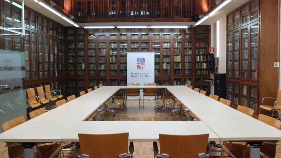 Interior de una sala de reuniones en la Universidad de Barcelona, con estanterías llenas de libros, mesas organizadas y sillas de madera.