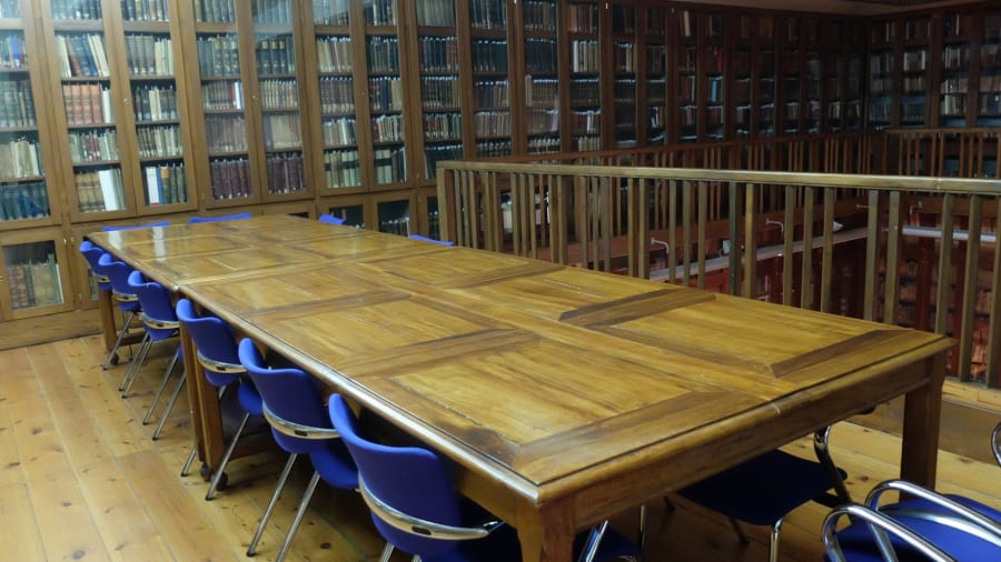 Interior de una biblioteca con estanterías llenas de libros y una mesa de madera rodeada de sillas azules.