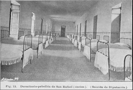 Dormitorio del pabellón de San Rafael con camas y mantas, mostrando un ambiente austero y vacío.