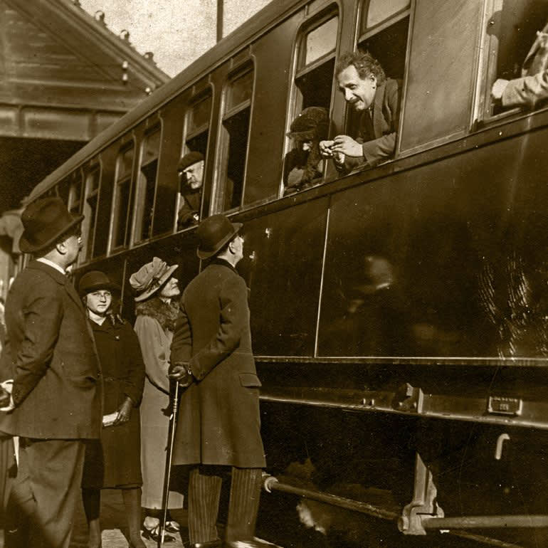 Albert Einstein asomado a la ventana de un tren, mientras un grupo de personas lo observa y conversa con él en una estación, en un ambiente de época.