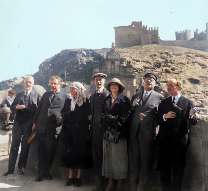 Grupo de personas posando en un lugar al aire libre, con una colina y una fortificación visible al fondo. Todos los miembros del grupo visten trajes formales y algunos utilizan sombreros. La escena parece ser un momento de alegría o celebración.