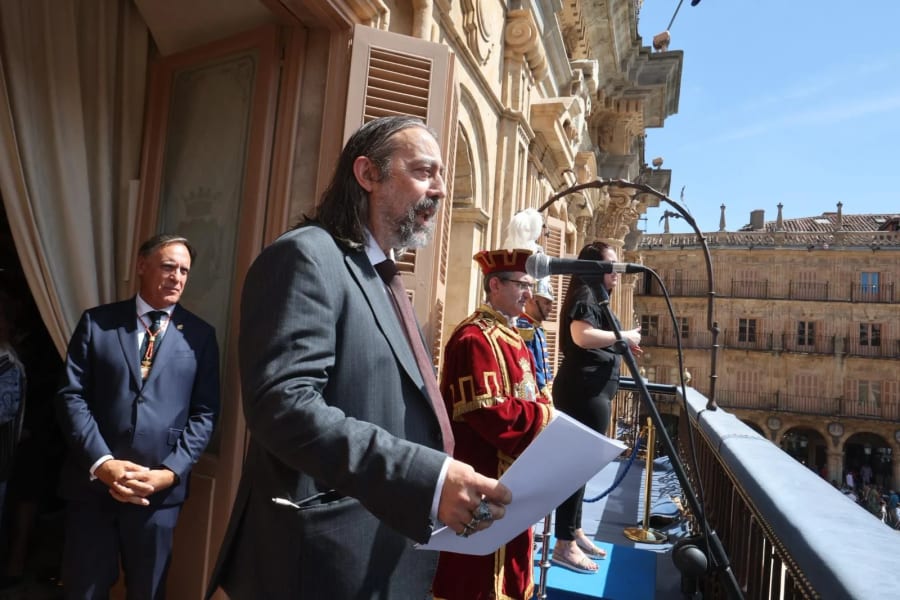 Una ceremonia en la que se ve a un hombre con barba y cabello largo, vestido de traje, sosteniendo papeles, mientras habla desde un balcón. A su lado hay una mujer con micrófono y un hombre vestido con un traje tradicional. En el fondo, se pueden ver edificios históricos.