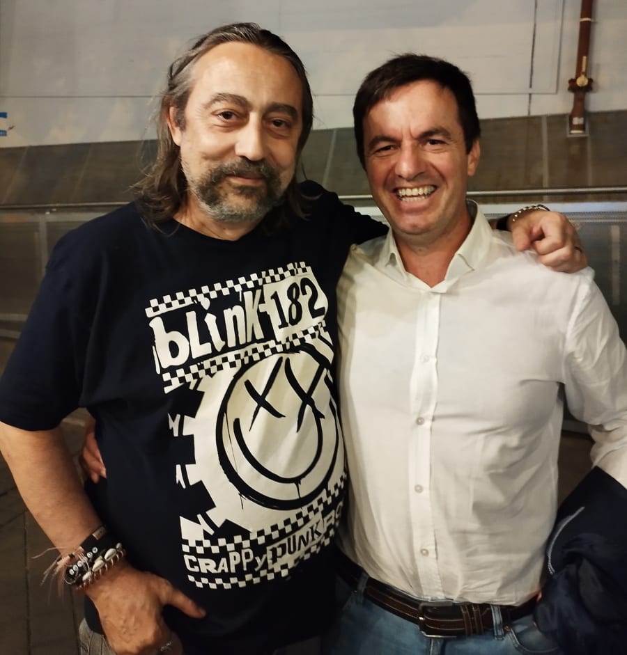 Dos hombres sonrientes posando juntos en un entorno informal. Uno lleva una camiseta negra con el logo de la banda Blink-182, mientras que el otro v viste una camisa blanca. El fondo muestra una pared y estructuras metálicas dentro de un lugar público.