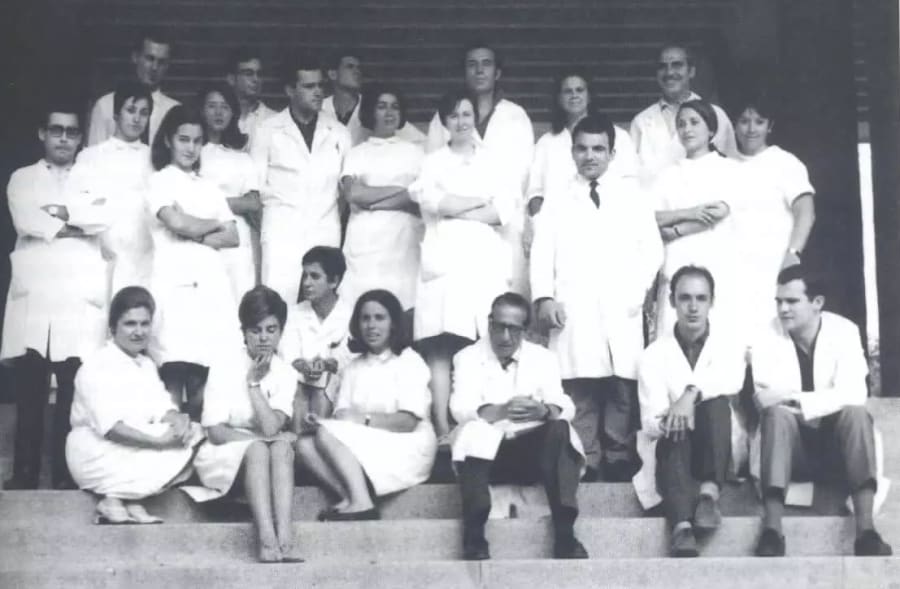 Grupo de científicos y estudiantes de medicina posando en el exterior de un edificio, todos vestidos con batas de laboratorio blancas, en una sesión de fotos grupal.