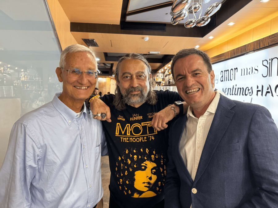 Tres hombres sonrientes posando juntos en un restaurante, uno viste una camiseta negra con un diseño amarillo, otro lleva una camisa blanca y el tercero usa una chaqueta azul.