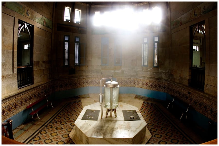 Interior del balneario de Aguas de Cabreiroá, mostrando una fuente octogonal de agua mineral con un diseño elegante, rodeada de paredes de piedra con ventanas y bancos de madera.