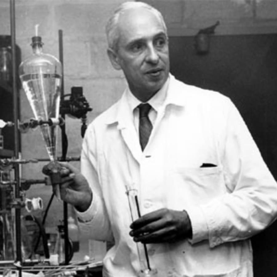 Un científico de pie en un laboratorio, vestido con una bata blanca, sosteniendo un frasco en una mano y un tubo de ensayo en la otra, con equipo de laboratorio visible de fondo.