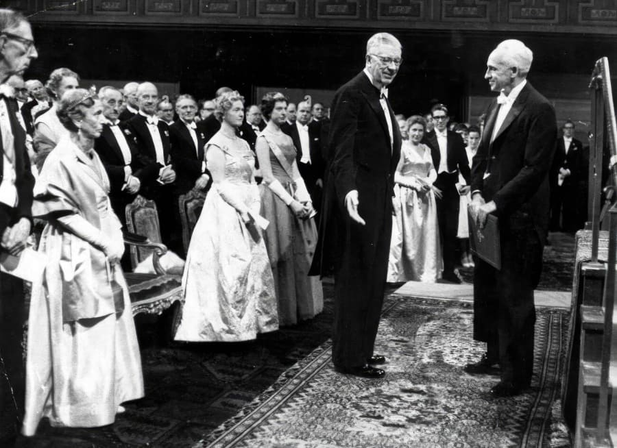 Ceremonia formal con un grupo de personas vestidas de gala, observando a dos hombres conversando en el centro de la habitación.