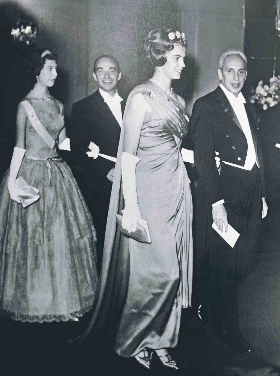 Una imagen en blanco y negro de un evento formal, donde varias personas están vestidas elegantemente. A la izquierda, una mujer lleva un vestido largo y una diadema, mientras que en el centro, un hombre sonríe. A la derecha, otra mujer con un vestido largo elegante se encuentra junto a un hombre vestido de gala.