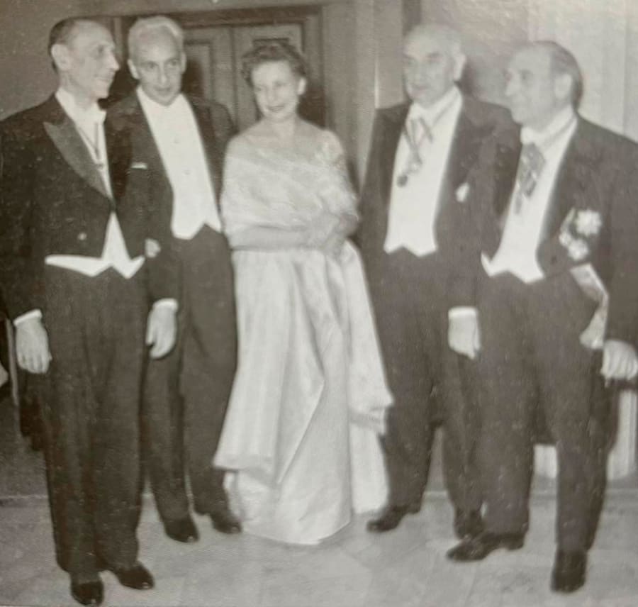 Cinco personas vestidas de gala posan para una fotografía en un evento formal, con un fondo interior elegante.