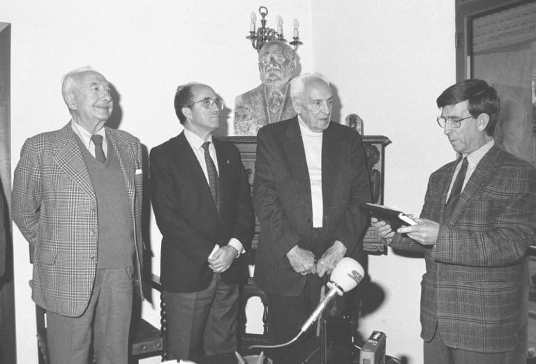 Cuatro hombres de pie en una habitación, con un busto de Santiago Ramón y Cajal visible detrás de ellos. Uno de los hombres está leyendo de un documento mientras los demás lo observan.