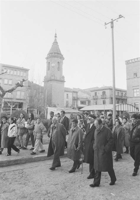 Una multitud camina por una calle en blanco y negro, con un reloj en una torre visible al fondo y edificios antiguos a los lados.