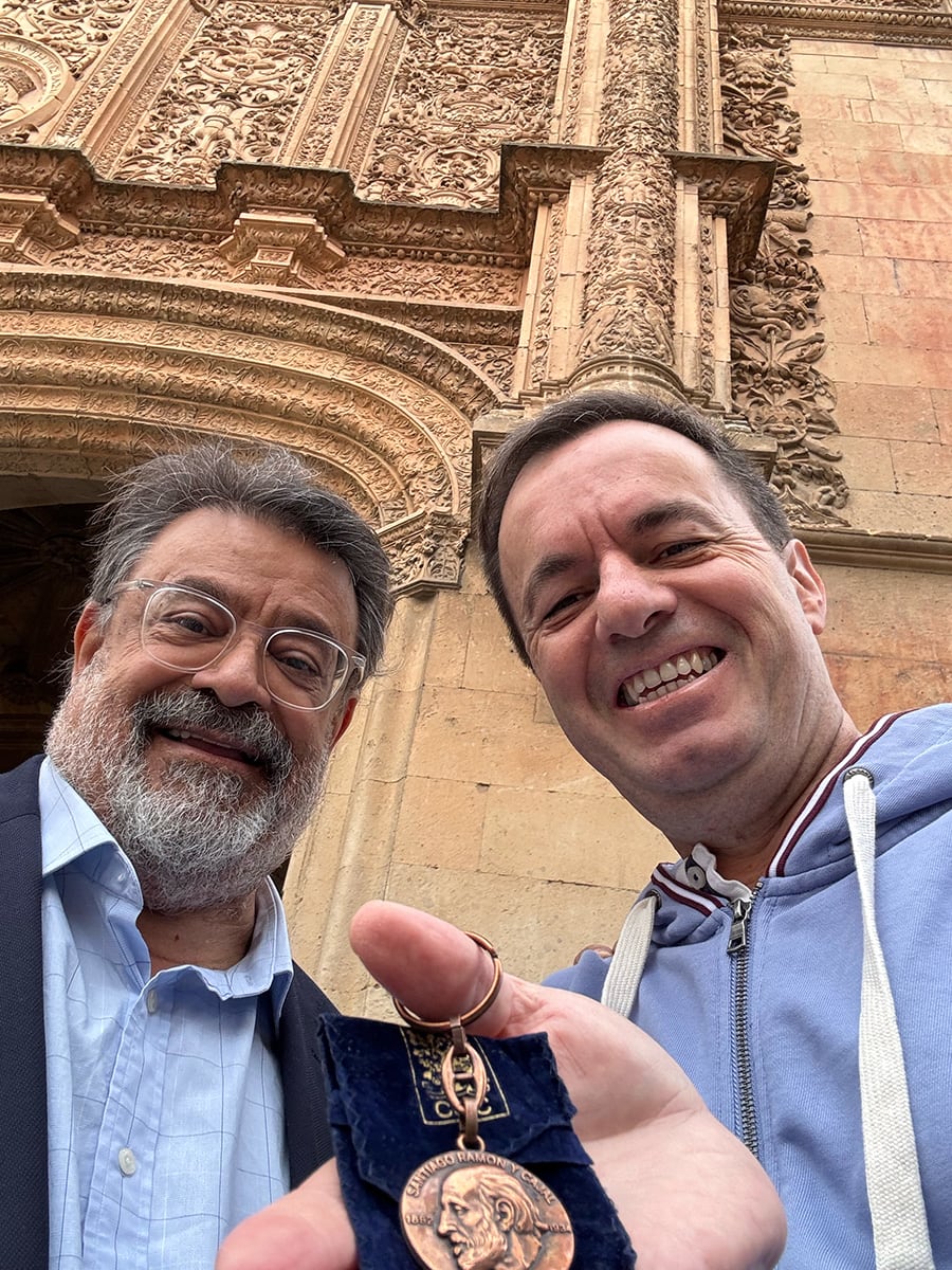 Dos hombres posando en primer plano frente a un fondo arquitectónico decorado, sosteniendo un llavero con el retrato de Santiago Ramón y Cajal.