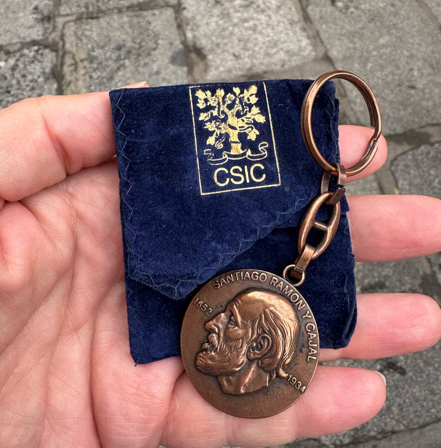Llave con medallón de Santiago Ramón y Cajal, sostenida en una mano, sobre fondo de pavimento. La medalla muestra un perfil del notable científico, y está contenida en una funda azul con el logo del CSIC.