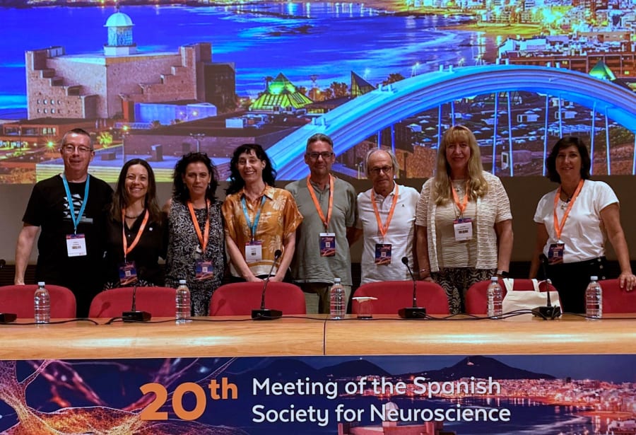 Grupo de personas posando en el escenario durante el 20º Encuentro de la Sociedad Española de Neurociencia, con un fondo que muestra una imagen iluminada de la ciudad.
