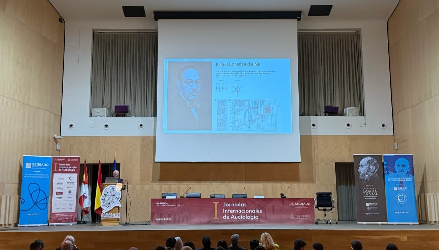 Un ponente presenta en el escenario durante las I Jornadas Internacionales de Audiología en Salamanca, con imágenes de Rafael Lorente de No en la pantalla y un panel de conferencias al fondo.
