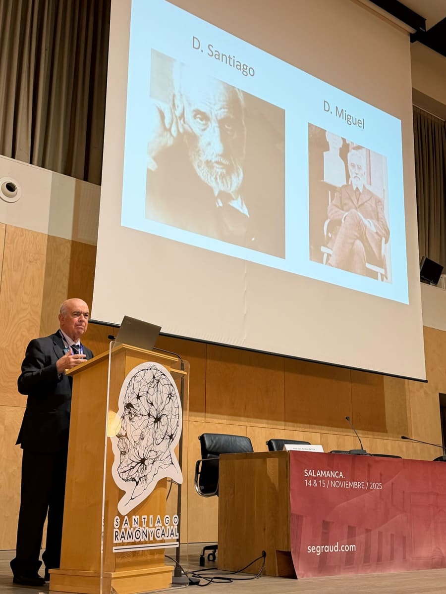 Un presentador en una conferencia está de pie frente a una pantalla que muestra imágenes de dos figuras históricas, D. Santiago y D. Miguel, con un diseño gráfico de un cerebro y la frase 