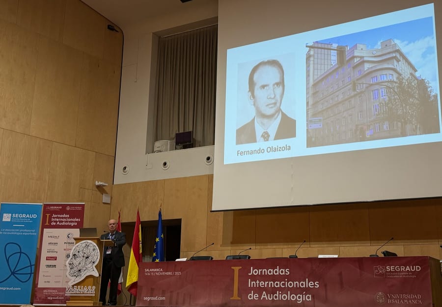 Un ponente presenta en el escenario durante las Jornadas Internacionales de Audiología en Salamanca, con una proyección de la imagen de Fernando Olaizola y un edificio en la pantalla de fondo.