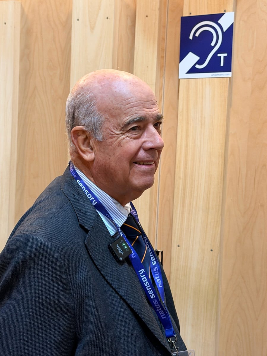 Retrato de un hombre mayor con cabello canoso y una chaqueta gris, sonriente, en un entorno de madera, junto a un signo que indica accesibilidad auditiva.