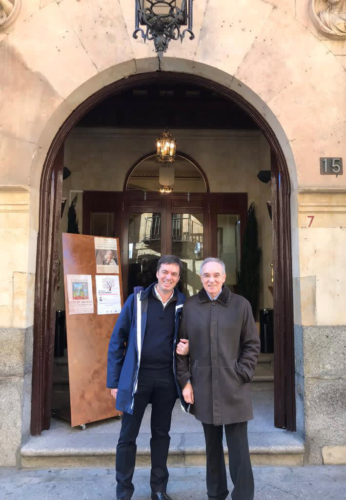 Dos hombres posando sonrientes frente a la entrada de un edificio histórico con un cartel visible en la pared.