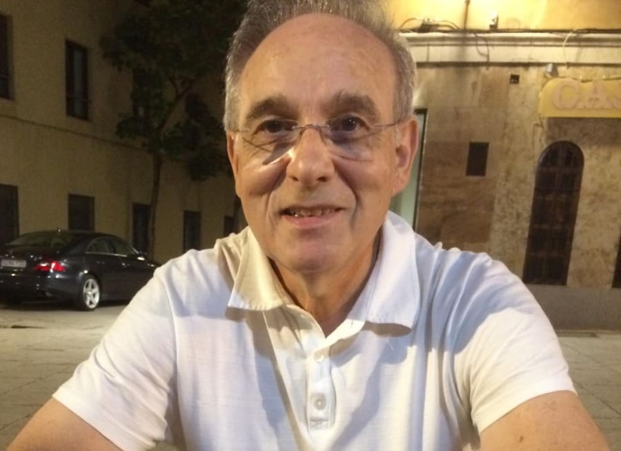 Retrato del Profesor José Luis López-Pérez sonriendo, vestido con una camiseta blanca, sentado en un entorno al aire libre con edificios de fondo.