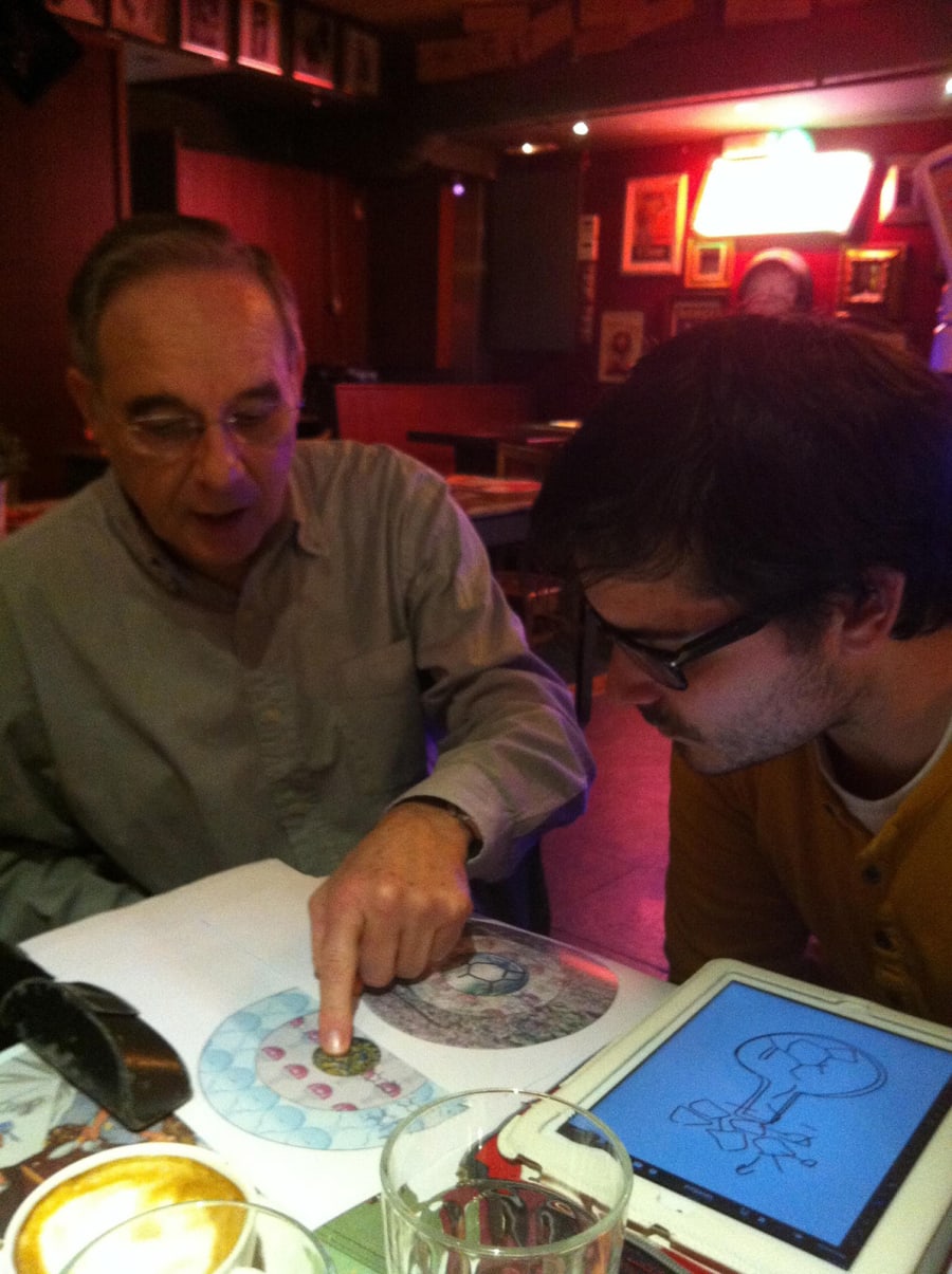 Dos personas sentadas en un entorno informal, discutiendo sobre gráficos científicos en una mesa, donde uno de ellos señala un diagrama en un papel mientras el otro observa con atención.